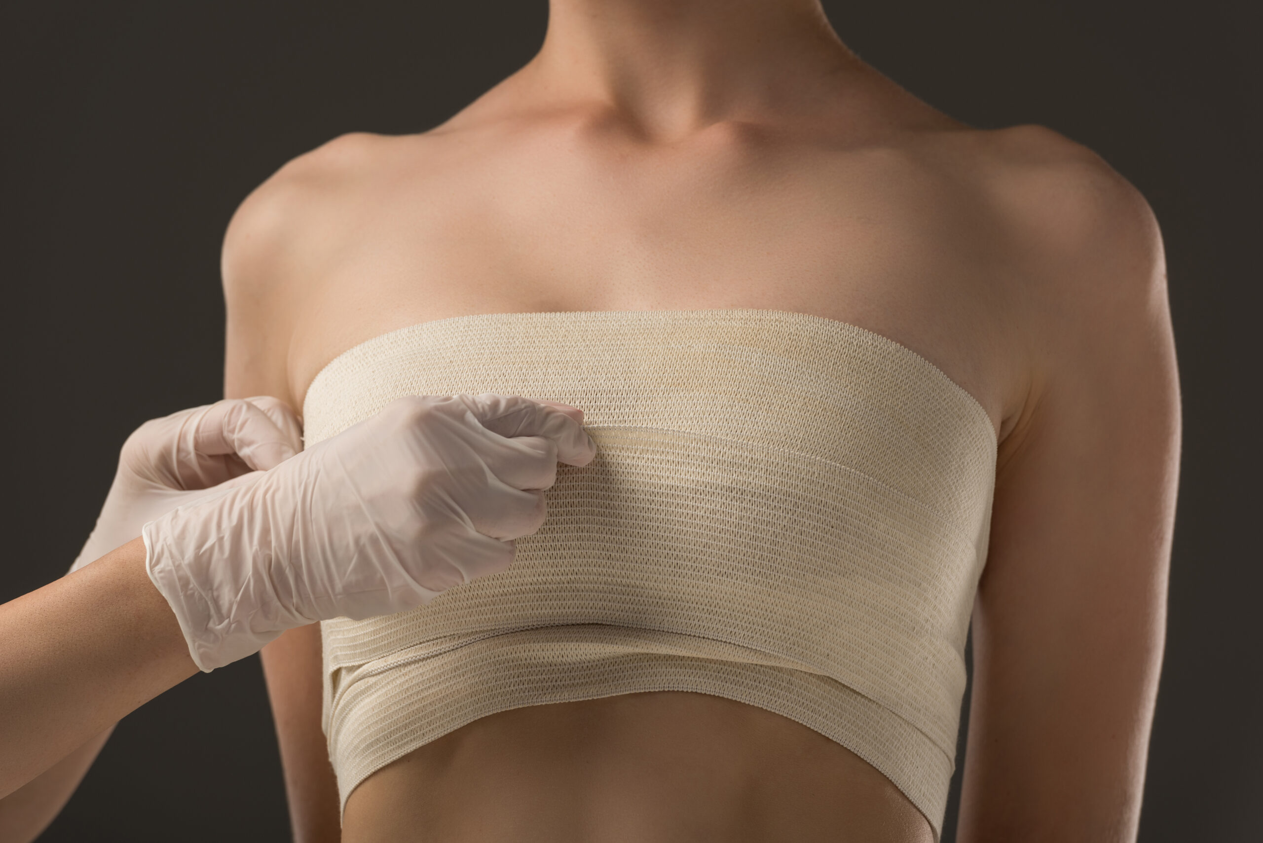 partial view of plastic surgeon in latex gloves and patient in breast bandage isolated on grey